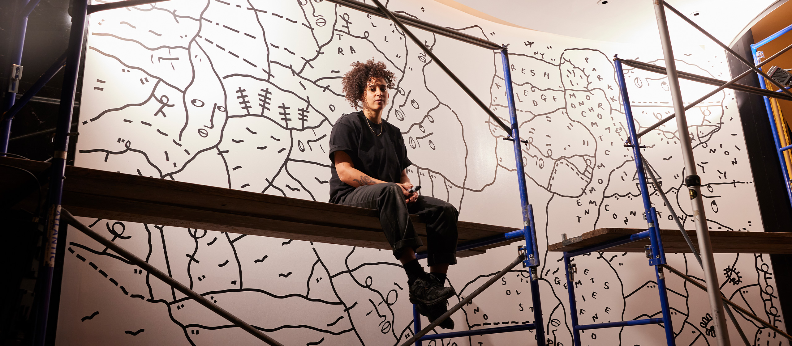 A person with curly hair sits on a scaffold, working on a large mural featuring abstract black lines and shapes on a white background. The mural covers a curved wall and scaffolding surrounds the person. They're wearing a black shirt and pants, focused on their work.