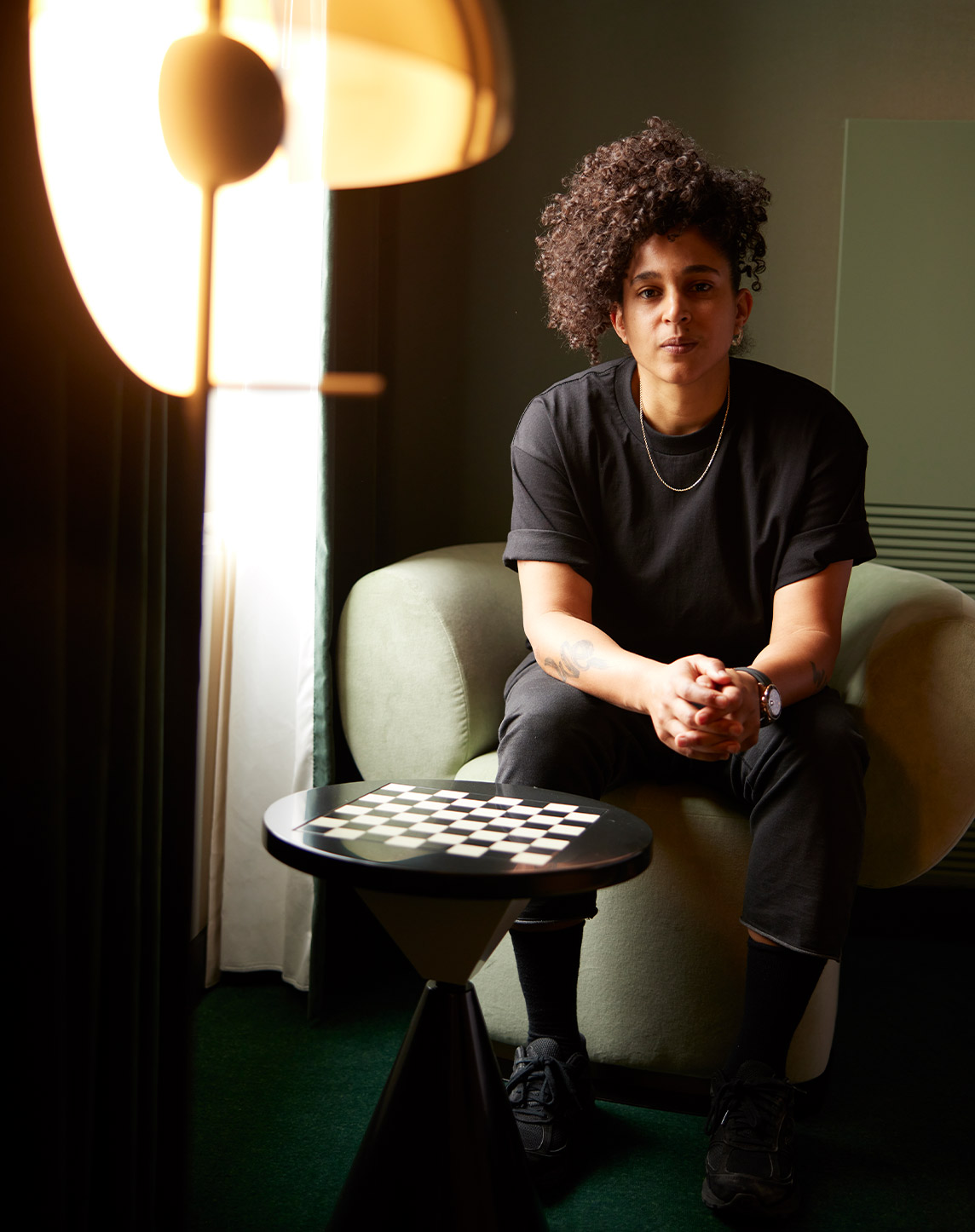 A person with curly hair sits thoughtfully on a green armchair, next to a small table with a checkered board, illuminated by soft light from a floor lamp.