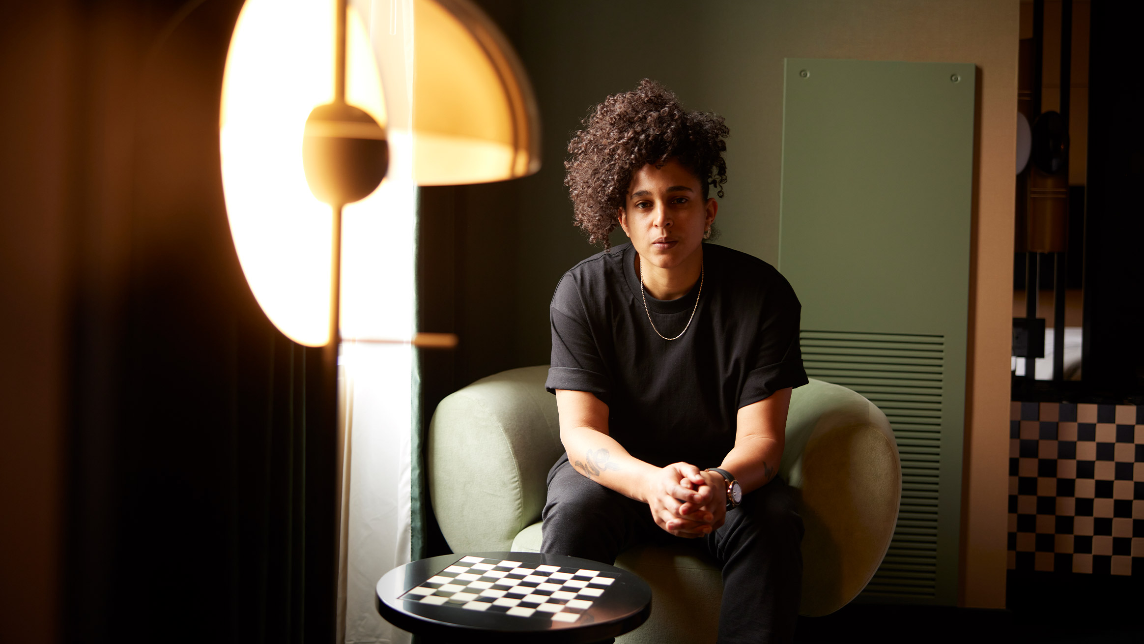 A person with curly hair sits confidently on a green sofa, facing the camera, in a stylishly decorated room with warm lighting and a modern chessboard on a table nearby.