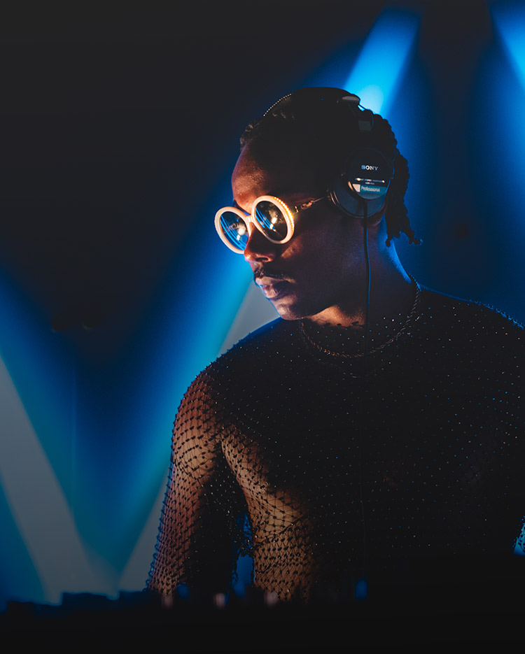 A dj wearing reflective round glasses and headphones focuses on mixing tracks, illuminated by blue stage lights, creating a vibrant atmosphere.