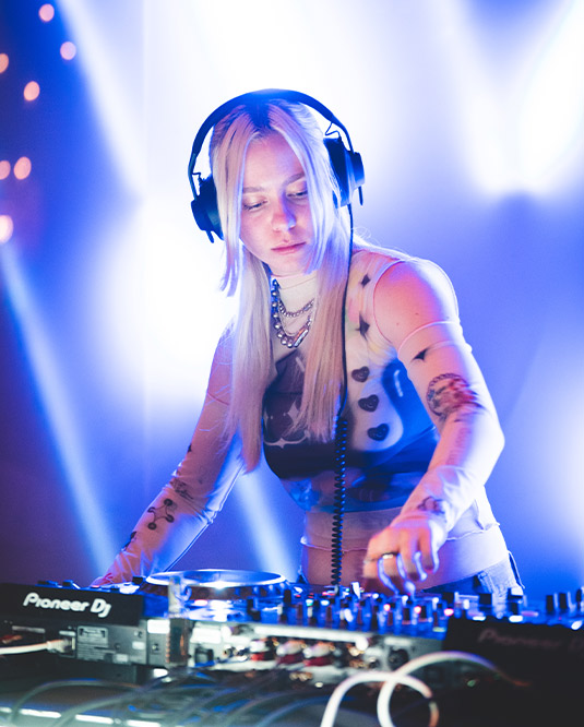 A female dj with long blonde hair, wearing headphones, is focused on mixing tracks on a pioneer dj console under vibrant stage lighting.