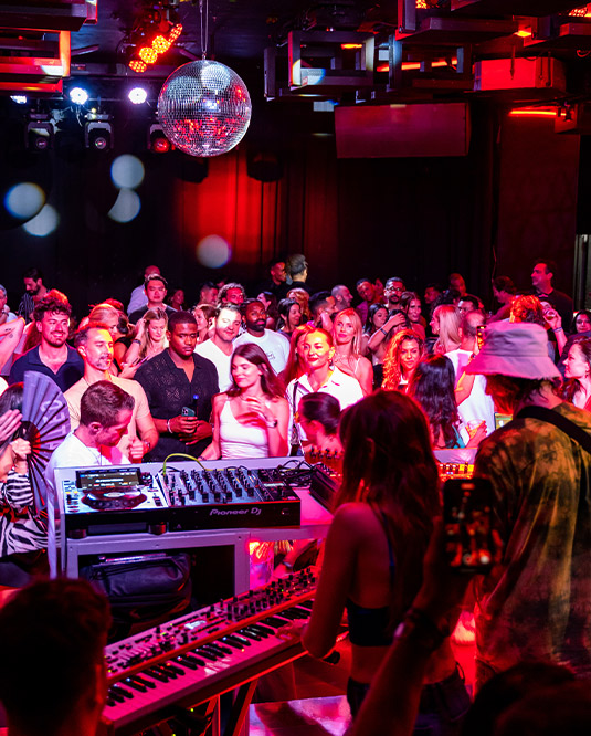 A vibrant nightclub scene with a diverse crowd watching a dj perform under a disco ball, illuminated by red and ambient lighting.