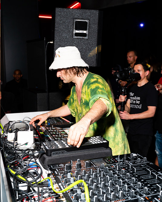 A dj in a tie-dye shirt manipulates equipment at a nightclub, surrounded by a crowd and dim, colorful lighting.
