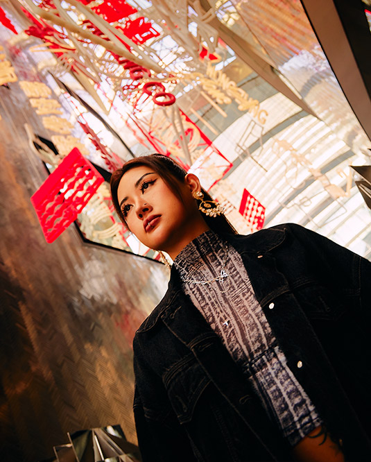 A woman in a denim jacket and large earrings looks pensively upwards, surrounded by abstract red neon sculptures and reflective surfaces.