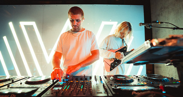 A male dj adjusts equipment while a female guitarist plays beside him, illuminated by vibrant vertical light bars in a moody, club-like setting.