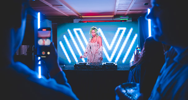 A dj with blonde hair, wearing headphones and a sequined outfit, performs at a club with blue neon lights and a vibrant 