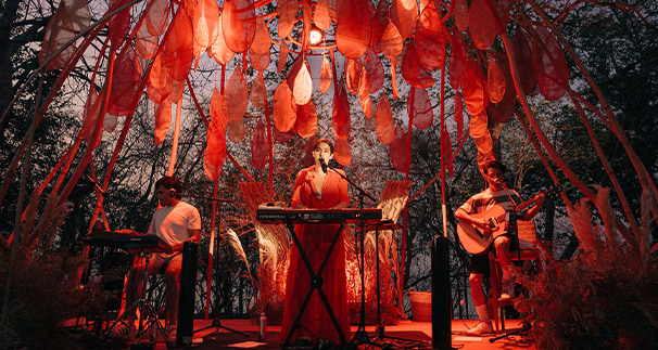 An enchanting outdoor music performance at night under a canopy of glowing red lanterns, featuring a vocalist in the center flanked by a keyboardist on the left and a guitarist on the right.