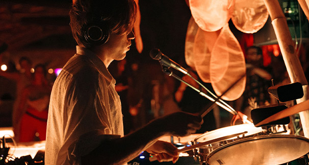 A young drummer wearing headphones performing at a dimly lit venue, illuminated by warm ambient lighting, intensely focused on playing a drum set.