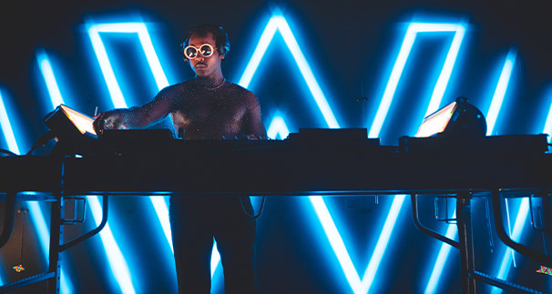 Dj in sunglasses and a shimmering top performs at a console, illuminated by bold blue neon lights forming triangular designs in the background.