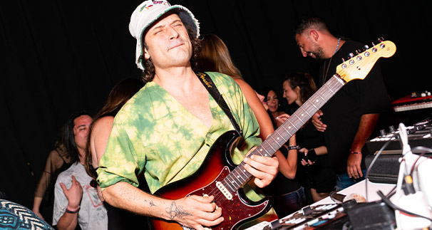 A man joyfully playing an electric guitar onstage, wearing a green tie-dye shirt and a white bucket hat, surrounded by an enthusiastic crowd.