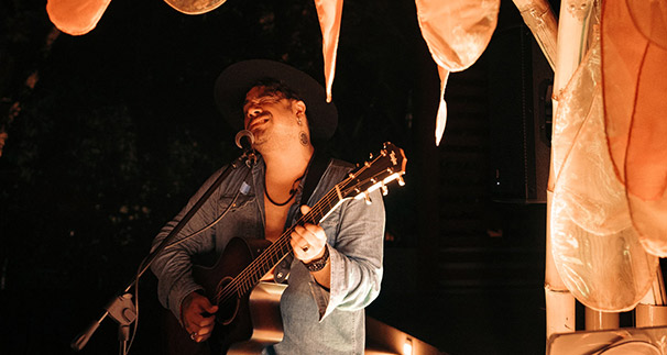 A musician in a hat playing an acoustic guitar at night, illuminated by warm ambient lights with soft, glowing lanterns in the background.