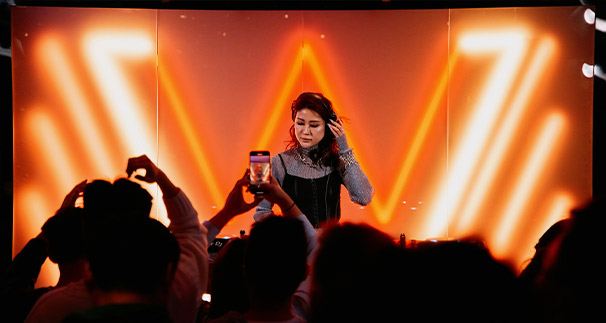 A female dj at a club, adjusting her headphones, with vibrant orange v-shaped lighting in the background. in the foreground, a crowd with raised hands, one holding a smartphone capturing the scene.