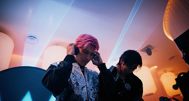 Two stylish men in a dimly lit room with modern lighting, one fixing his pink hair, and the other looking down, both dressed in elaborate outfits.
