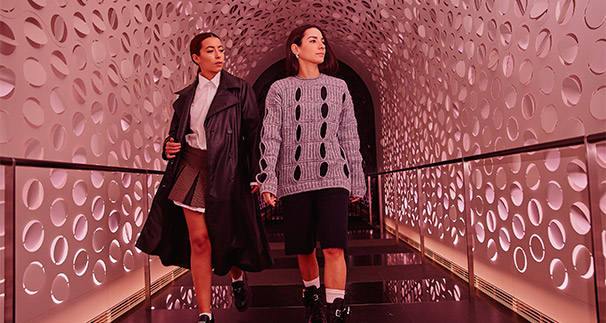 Two women walking through a futuristic tunnel with intricate wall patterns, one in a gray sweater and skirt, the other in a black coat and pants, illuminated by pink lighting.