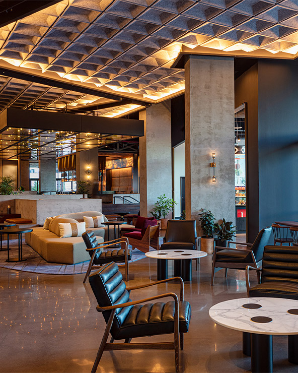 Modern hotel lobby with various seating arrangements, geometric ceiling design, and warm lighting, leading to a visible street view.