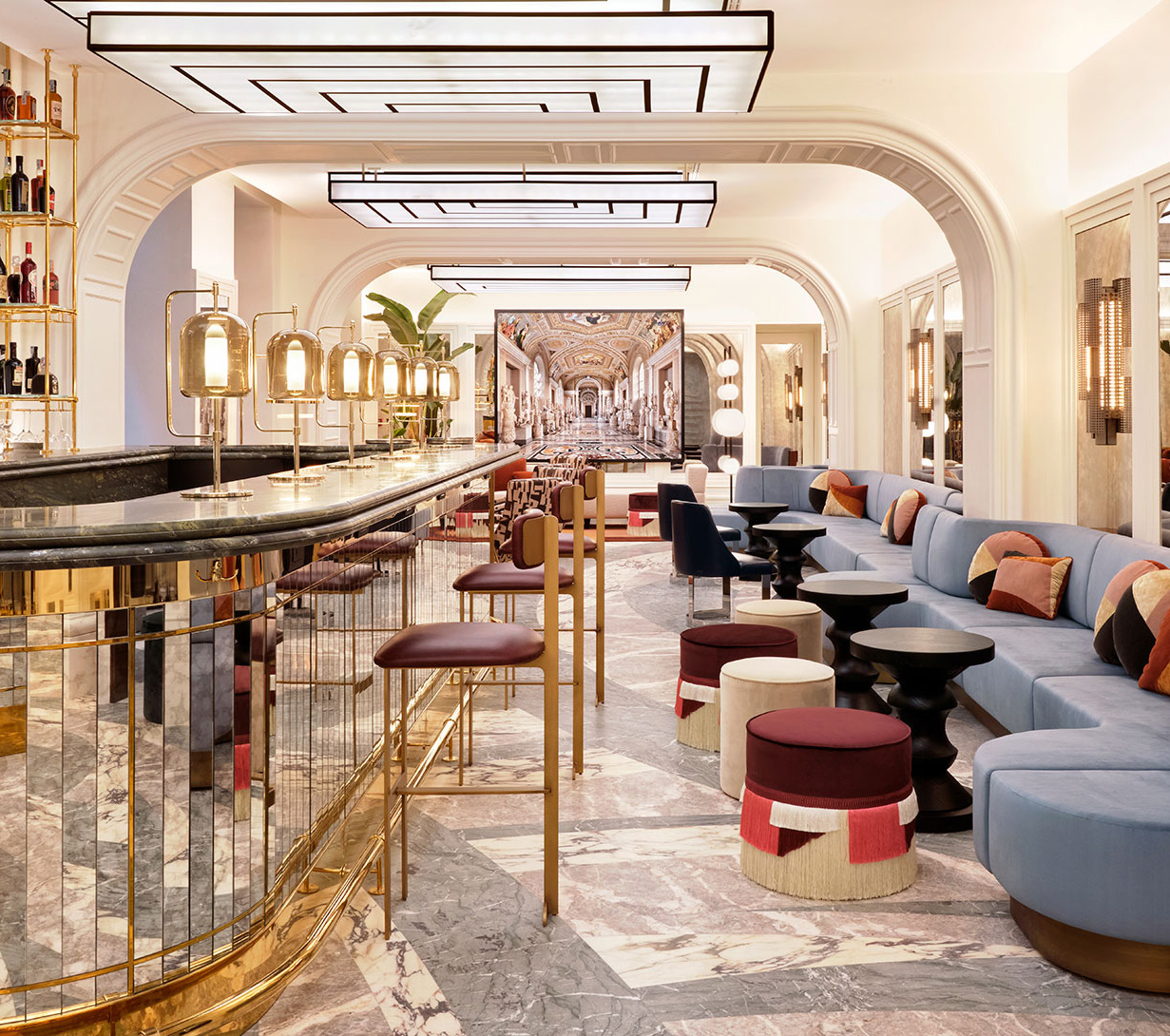 An elegant bar with a marble floor, plush seating in blue and crimson, and gold accents throughout. a well-lit ceiling and extensive shelving with bottles complete the luxurious setting.