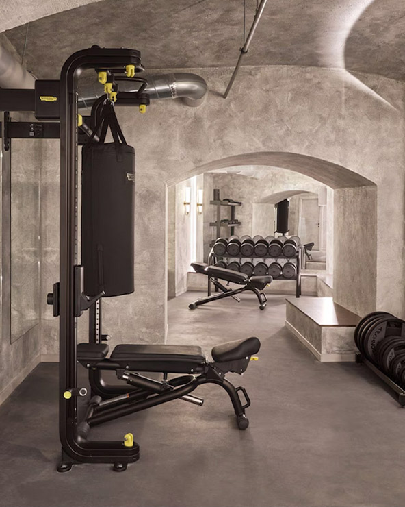 An elegantly designed gym with textured gray walls and archways, featuring a multi-station weight machine, a set of dumbbells, and mirrored walls.