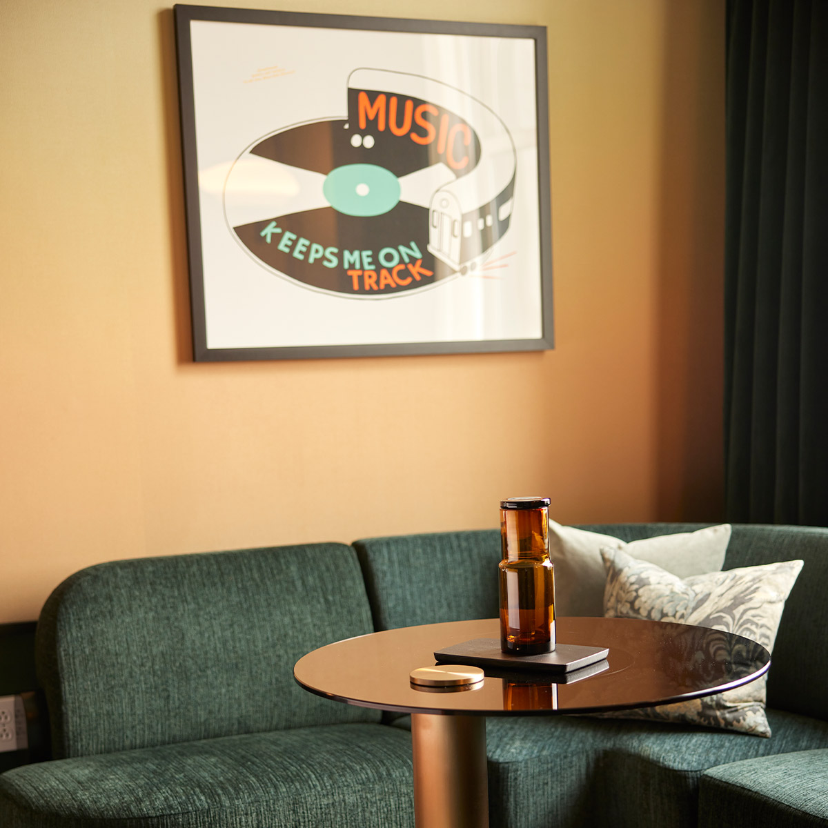 A cozy room corner featuring a small round table with a brown bottle and an open book, a green sofa with a pillow, and a framed poster proclaiming 