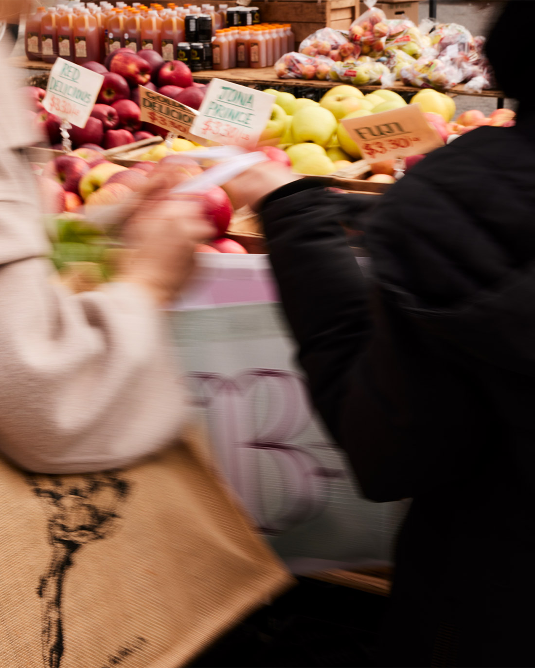 A customer at a bustling market stall, bag in hand, selects fresh fruit, with a variety of colorful produce and price signs in the background.