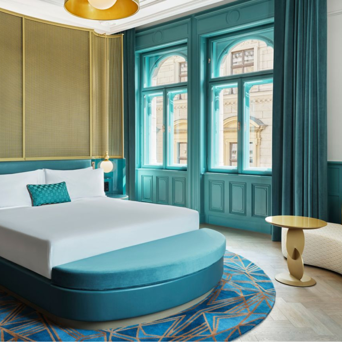 Elegant hotel room with teal walls and matching round bed, large windows with street view, golden ceiling light, and a small modern side table.