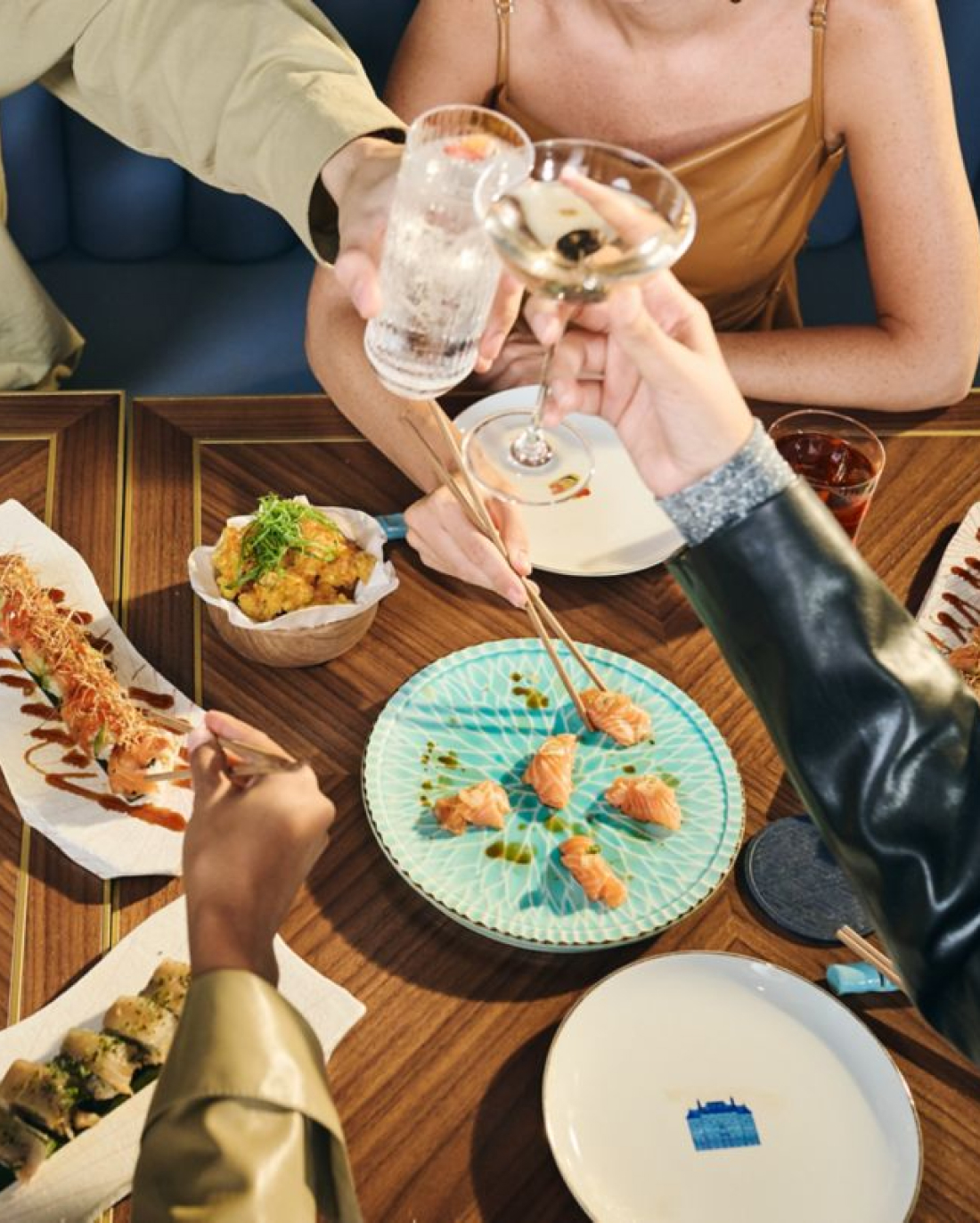 Three people cheers with drinks over a table featuring an array of sushi and appetizers in a cozy dining setup.