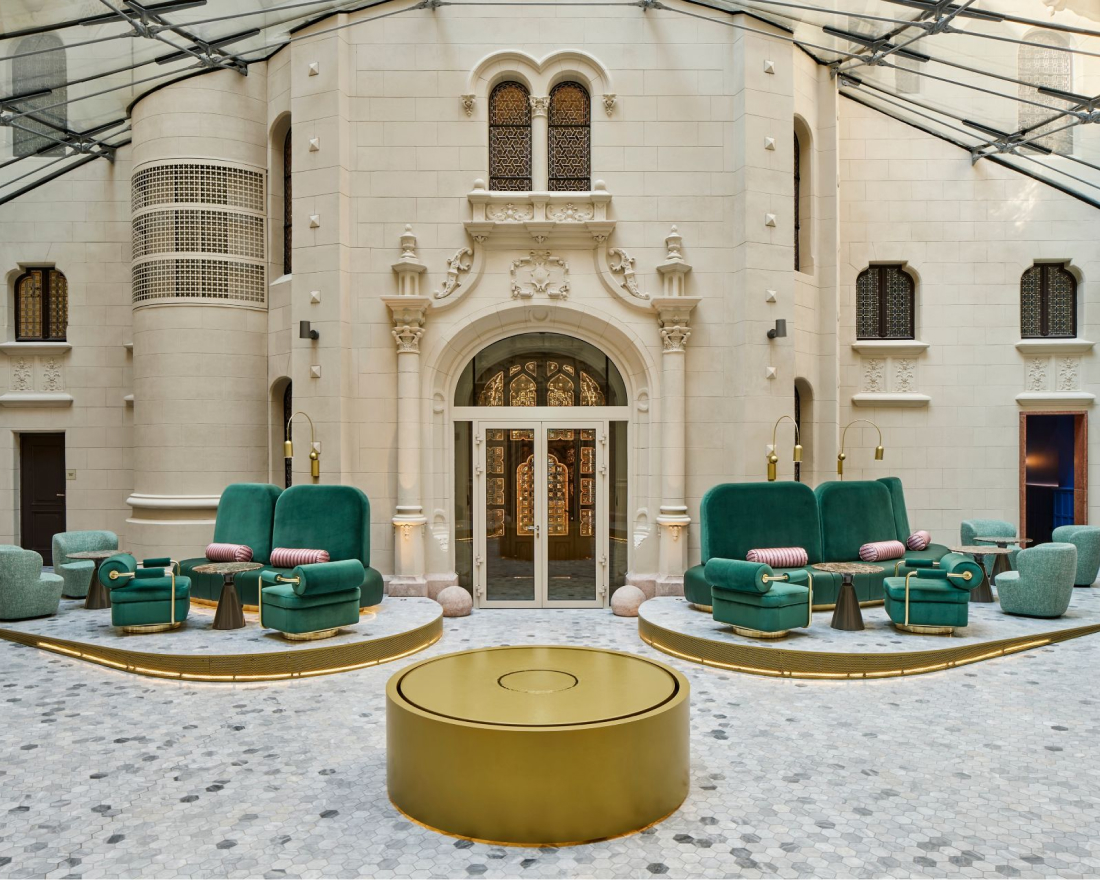 Luxurious hotel lobby with green velvet armchairs and a large circular gold coffee table, set against ornate white walls and arching doors under a translucent arched ceiling.