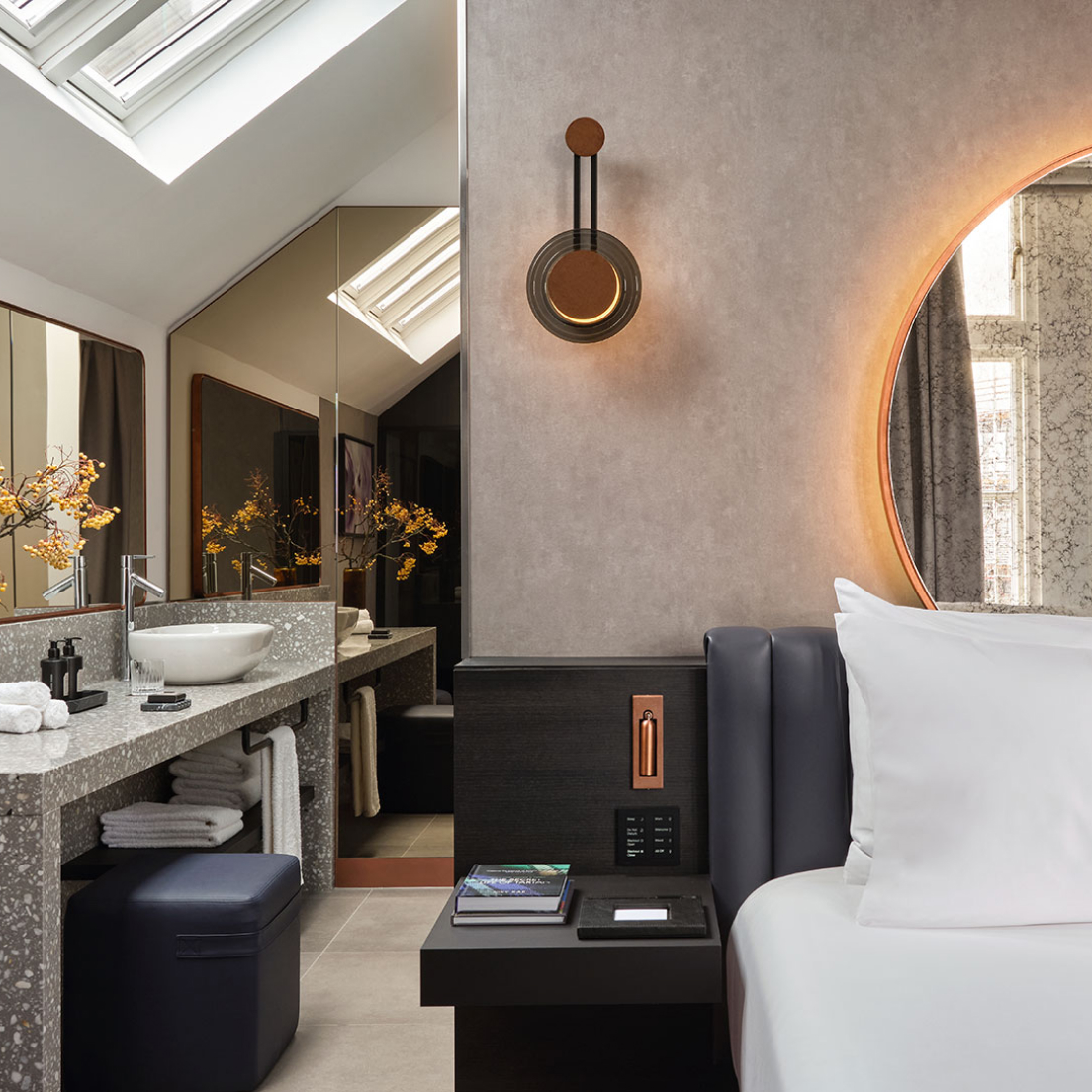 A modern hotel room featuring a neatly made bed with white linens next to a stylish bathroom area. the bathroom has a large mirror, a vanity with two sinks, and a terrazzo counter. ambient lighting enhances the cozy atmosphere.