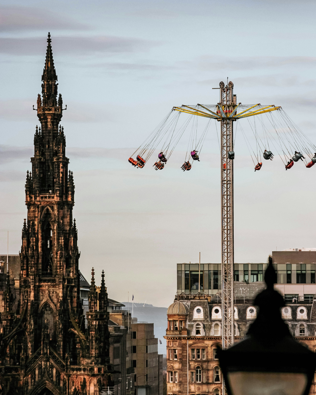 A towering swing ride spins against a cityscape featuring a gothic spire and modern buildings in reddish evening light.