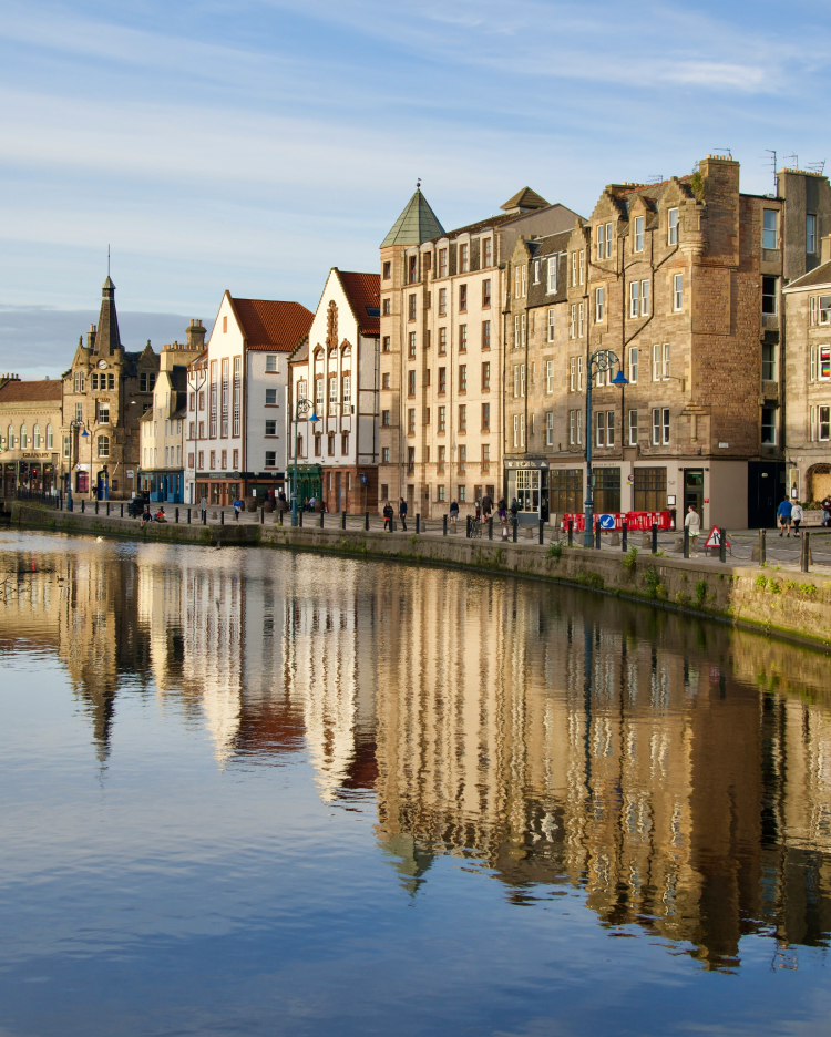 Historic buildings line the sunlit banks of a calm river reflecting the clear blue sky in a tranquil urban scene, with pedestrians walking along the riverfront in Edinburgh.