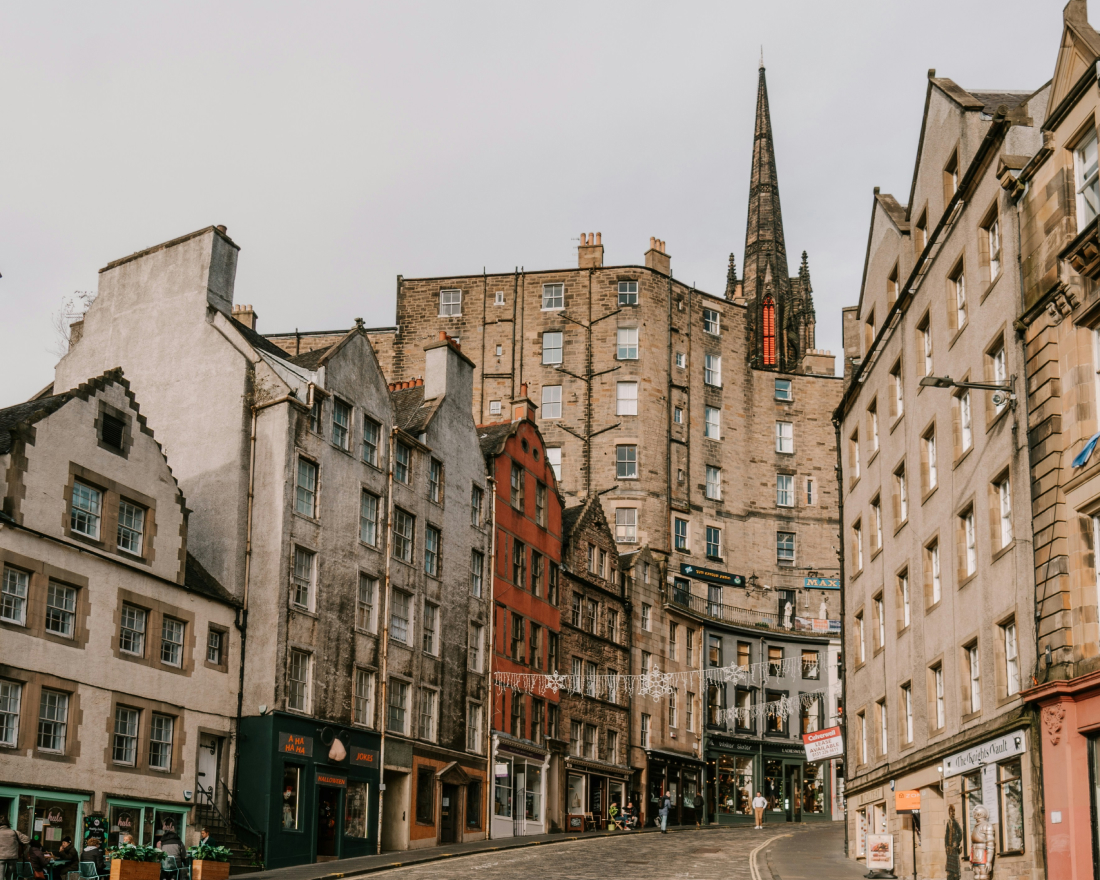 Historic street view in Edinburgh with old multistory buildings in various colors, a cobblestone road, and a distant gothic spire visible under a cloudy sky.