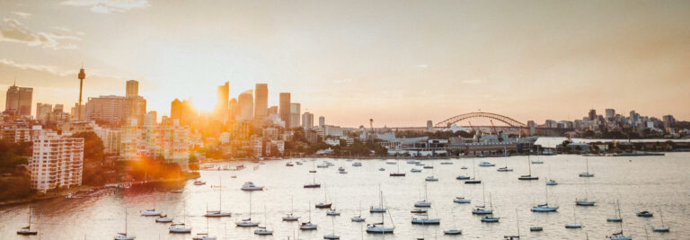 Panoramic view of sydney skyline at sunset, featuring silhouettes of skyscrapers, the sydney harbour bridge, and boats in the foreground on calm water.