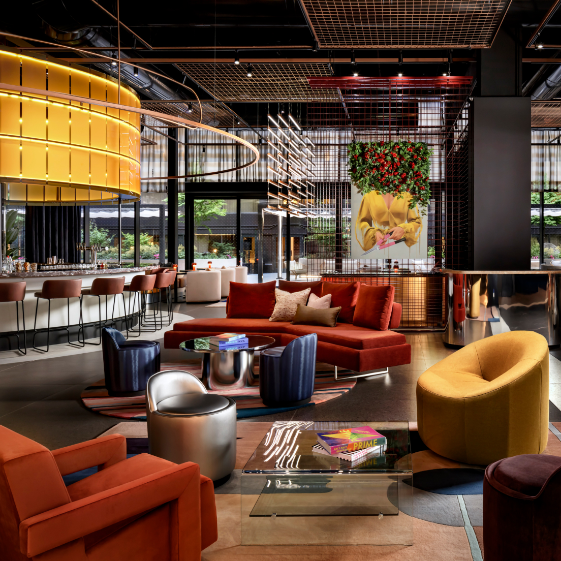 A stylish modern lounge with a bar area, featuring vibrant orange and blue seating, metallic accents, and a large flower arrangement hanging above a relaxed seating area.