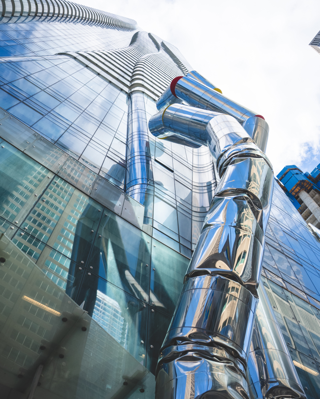 Low-angle view of a shiny, tubular stainless steel sculpture resembling a bent pipe installed along the exterior of a modern glass skyscraper with reflections of the sky and surrounding buildings.