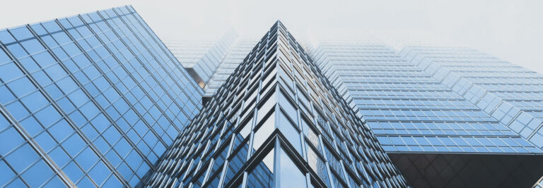 Upward view of a striking architectural convergence of several modern skyscrapers with reflective glass facades against a hazy sky, emphasizing geometric lines and angles.