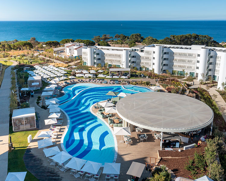 Aerial view of a luxury resort featuring a large, curved swimming pool with blue and white patterns, surrounded by loungers and white parasols. Modern white buildings and lush greenery surround the area. The clear blue sea is visible in the background under a bright, sunny sky.