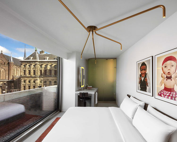 A minimalist hotel room features a large bed with white linens, a modern light fixture, and two playful portraits on the wall. A floor-to-ceiling window offers a view of historic buildings outside. The room includes a small desk and a frosted glass bathroom area.