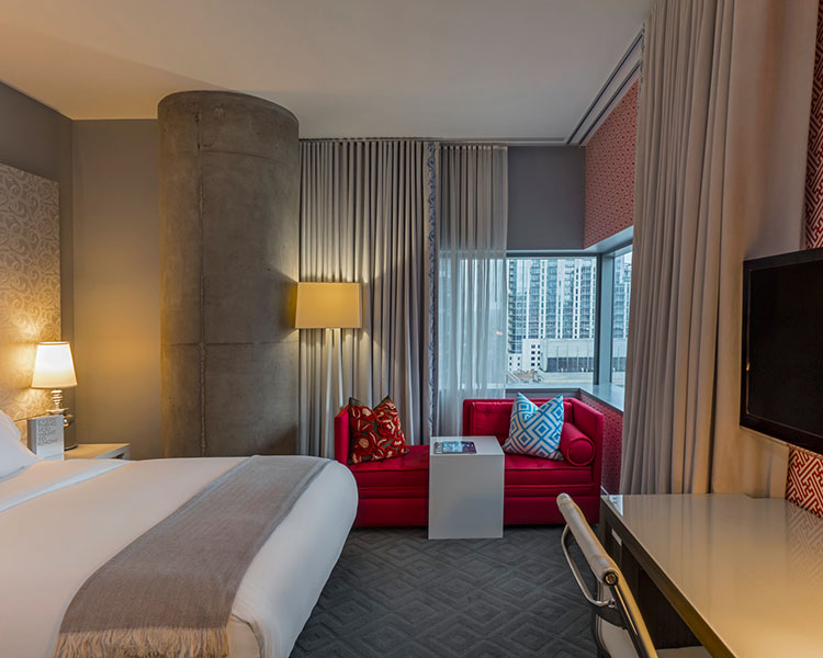 A modern hotel room featuring a neatly made bed with white linens and a beige throw, a red sofa adorned with colorful pillows, a desk with a chair, and a large window with a city view. The room has a soft, ambient lighting and a contemporary decor.