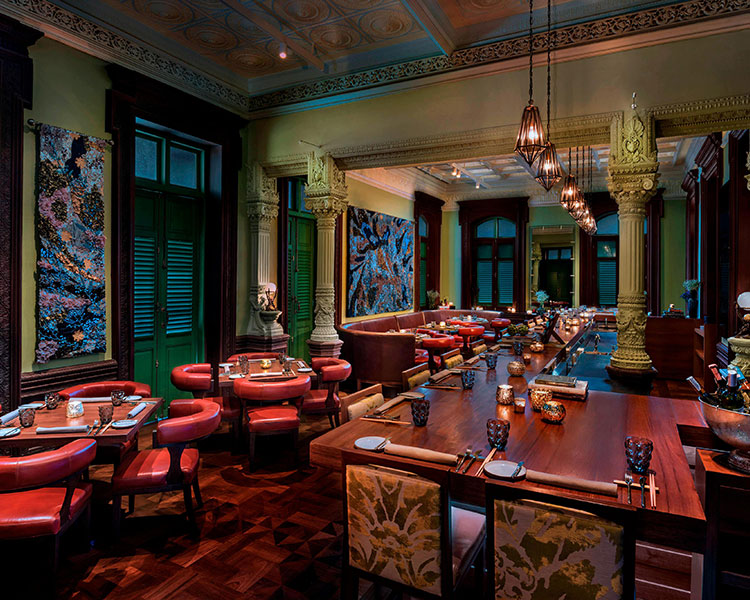 A cozy, upscale dining room with a blend of classic and modern decor. There are red cushioned chairs around wooden tables, ornate pillars, pendant lighting, and vibrant artwork on the walls. The room features a warm, inviting ambiance with delicate lighting and intricate ceiling designs.