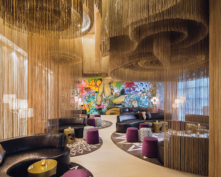A modern lounge features circular seating areas with black sofas and purple stools, separated by cascading string dividers. The walls display vibrant graffiti art, and small golden round tables are placed throughout, creating an eclectic and artistic ambiance.