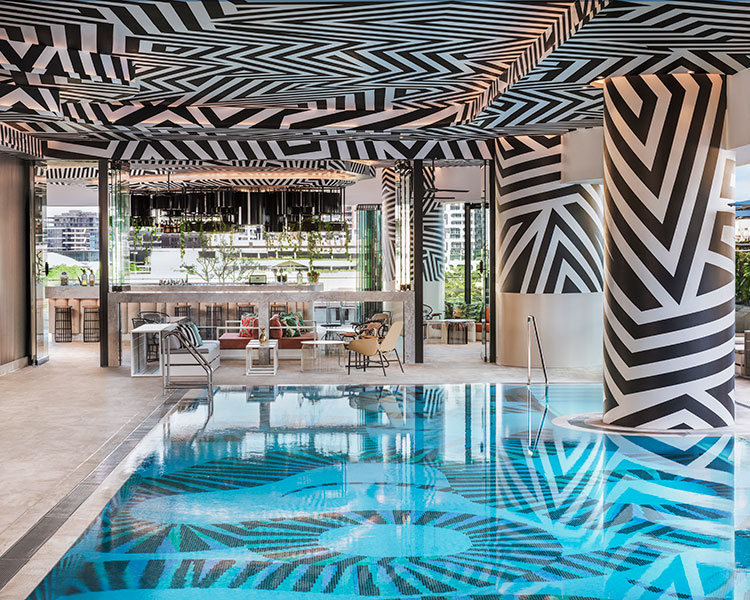 A modern indoor pool area with zebra-stripe inspired black and white patterns covering the walls, ceiling, and columns. The pool reflects these patterns, creating a mesmerizing visual effect. In the background, there are lounge areas with chairs and tables.