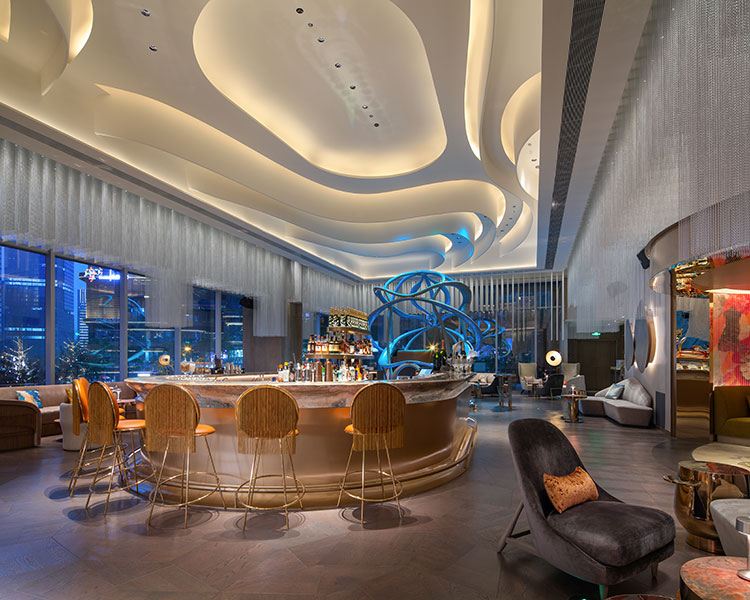 A modern and stylish bar interior with a sculptural ceiling and ambient lighting. The circular bar has elegant bar stools around it, and there is a unique blue spiral structure in the background. The seating is luxurious, with a mix of chairs and sofas.