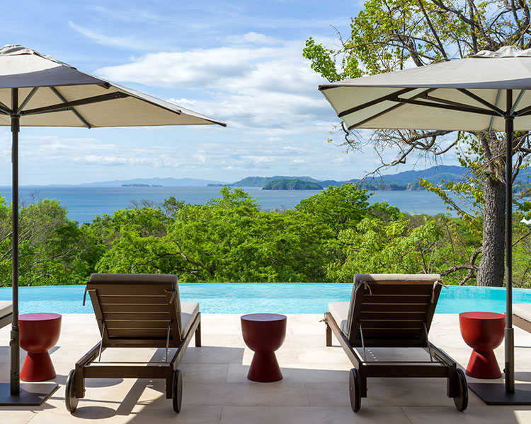 Two poolside lounge chairs under large umbrellas overlooking a scenic infinity pool. In the background, there is lush greenery and a view of the ocean and distant islands under a partly cloudy sky. Red side tables are placed between the chairs.