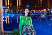 A woman in a vibrant green top and a colorful skirt stands by a lit pool at night. The background is filled with illuminated buildings and blurred figures, creating a lively, urban atmosphere. The scene has an upscale, festive vibe.