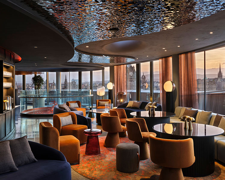 A modern, stylish lounge with a large window view of an urban cityscape during sunset. The room features plush chairs and sofas in warm tones, round tables, and contemporary decor. A reflective ceiling and ambient lighting enhance the cozy, upscale atmosphere.