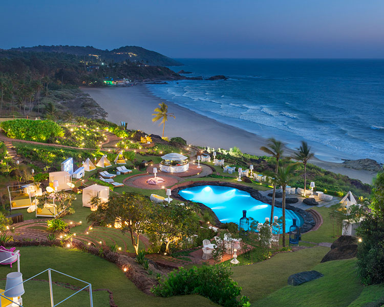 A scenic view of a coastal resort at dusk. The resort features a lit pool, surrounding greenery, and several lounge areas and cabanas. Beyond the resort, waves gently lap at the sandy beach, and hilly terrain is visible in the background.