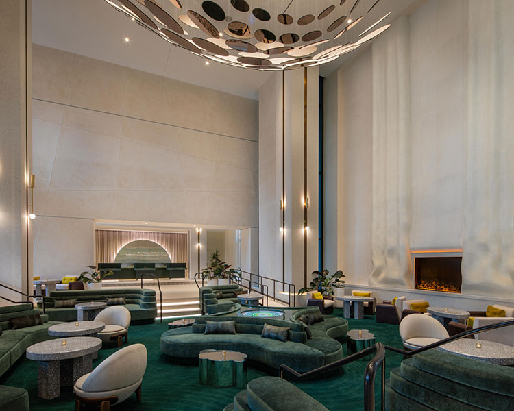 A modern, luxurious hotel lobby with high ceilings and minimalist decor. The space features green velvet seating, round tables, a large, abstract ceiling light fixture, and a tall fireplace with a broad, flowing backdrop. A front desk is visible in the background.
