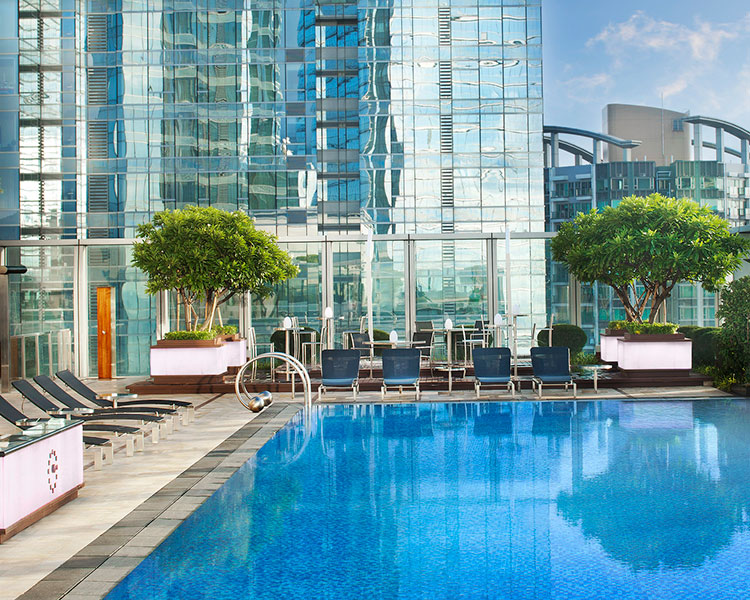 A luxurious outdoor pool area in a modern urban setting. The pool is surrounded by lounge chairs, shaded seating areas, and potted plants. The tall glass buildings in the background reflect the bright blue sky, creating a serene and refreshing atmosphere.