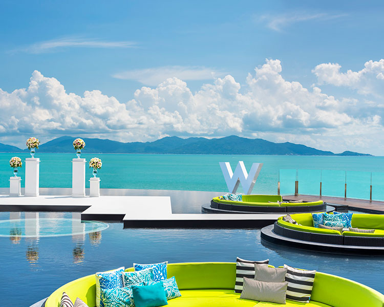 A scenic view of a coastal resort featuring round, green seating areas surrounded by water. The background showcases a calm turquoise sea, distant mountains, and a clear blue sky with fluffy clouds. Several flower arrangements and a large 