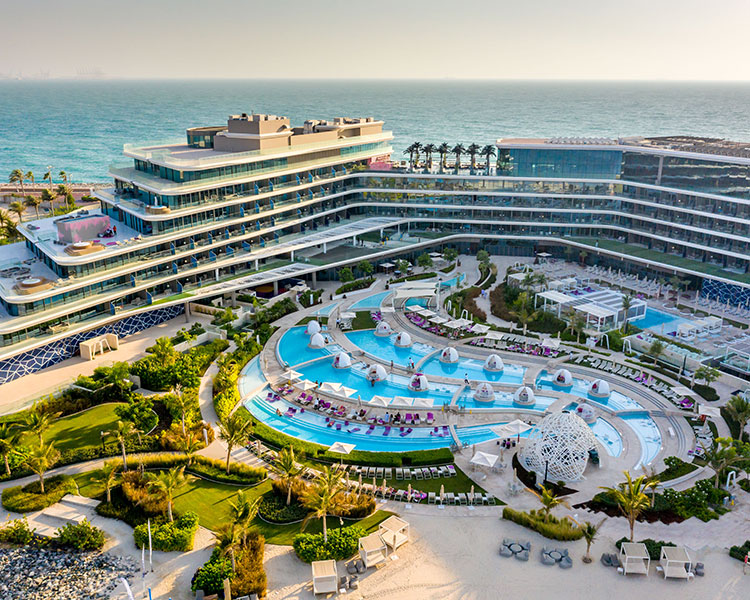 Aerial view of a luxurious beachfront resort with multi-level buildings overlooking a large, winding swimming pool. The pool area is surrounded by lounge chairs, cabanas, and landscaped gardens. The resort faces a calm, expansive sea under a clear sky.