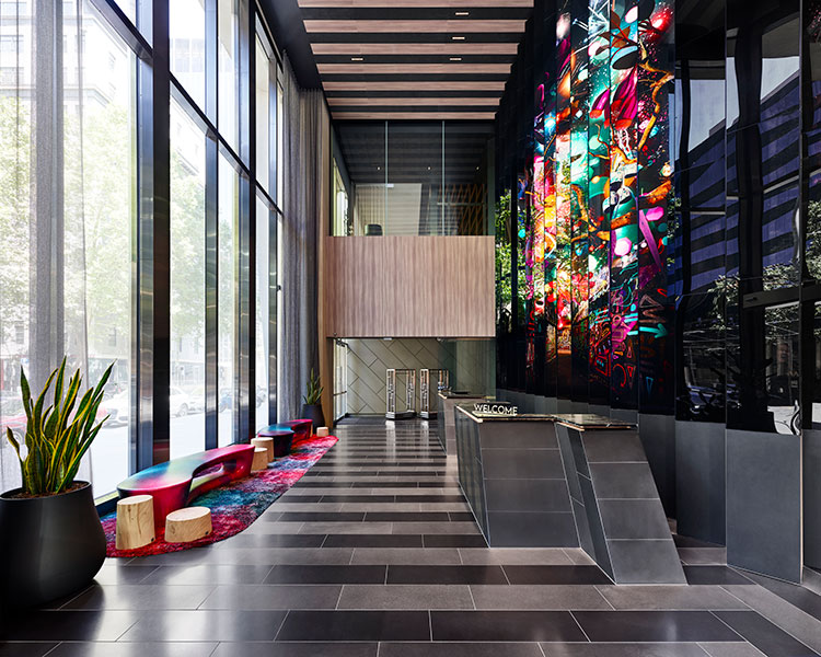 A modern lobby with floor-to-ceiling windows, a sleek reception desk, and colorful abstract wall art. The space has contemporary furnishings, including a long wooden bench with cushions and a potted plant. Natural light brightens the room through large windows.